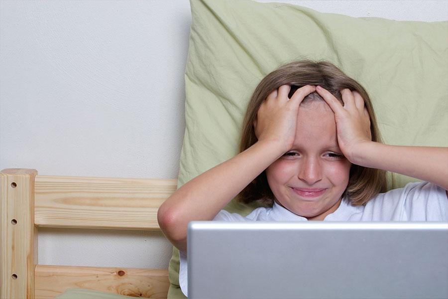 mała dziewczyna siedzi na drewnianym łóżku przed laptopem trzymając ręce blisko twarzy