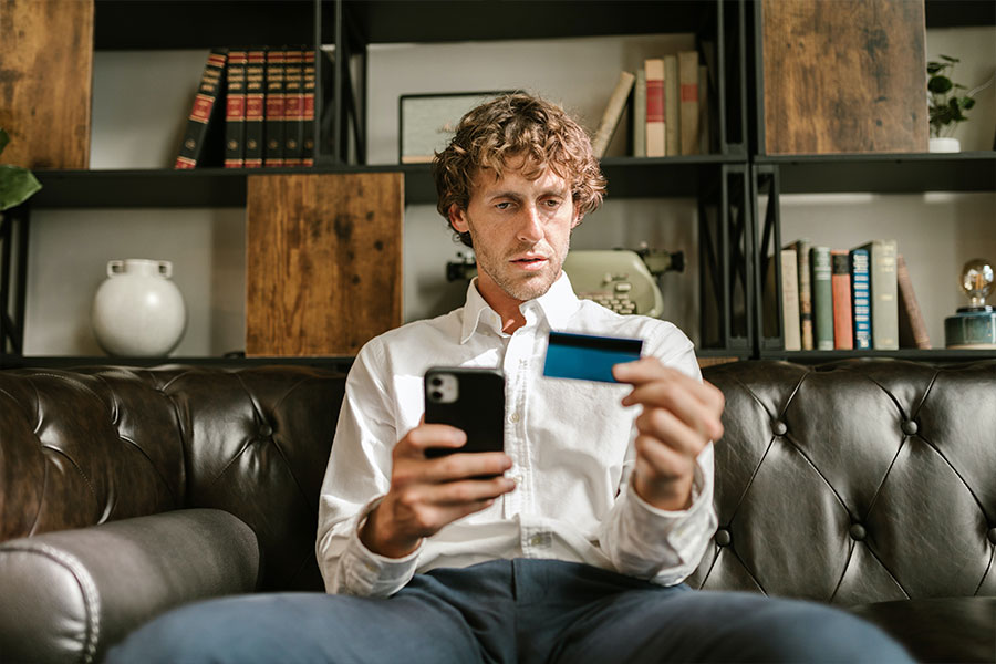 mężczyzna siedząc na kanapie patrzy w telefon i na kartę płatniczą