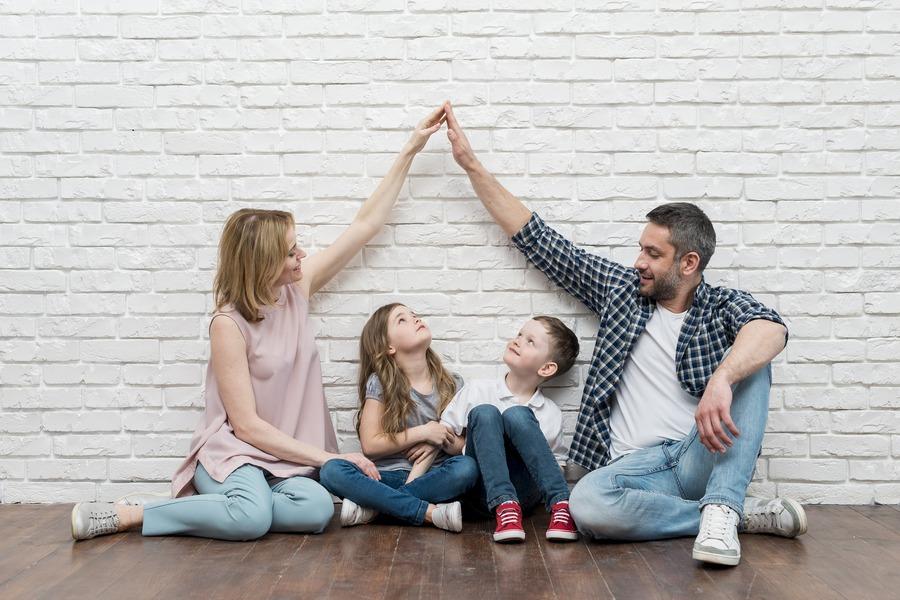 Rodzina zastępcza siedzi na podłodze: adoptowane dzieci między rodzicami. Para symbolicznie składa ręce w dach nad głowami pociech