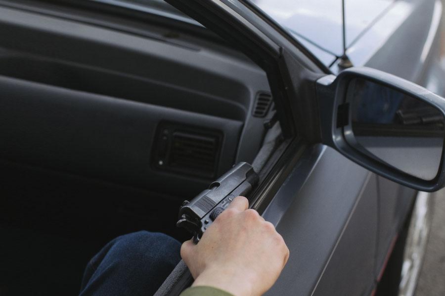 osoba siedzi w ciemnym samochodzie i trzyma pistolet w prawej ręce