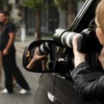 detektyw siedzi w ciemnym samochodzie w czarnej kurtce i robi zdjęcia pary