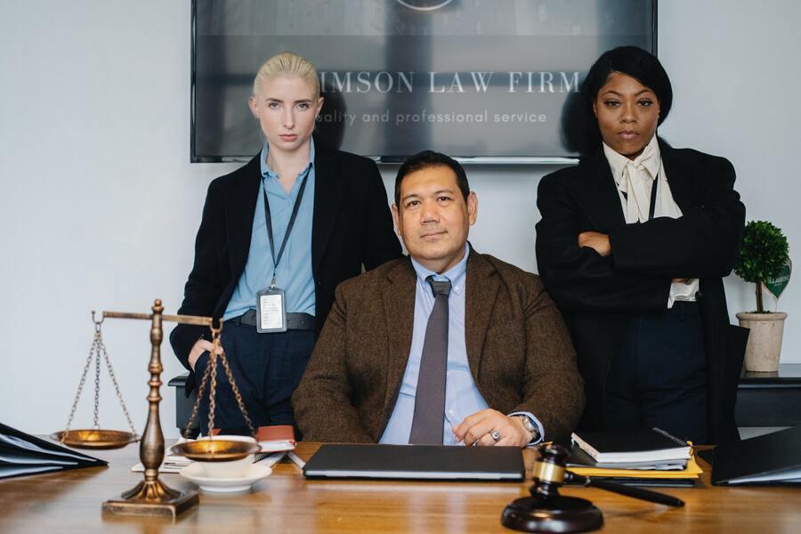 Prawnicy w eleganckich strojach. Mężczyzna w garniturze siedzi za biurkiem, między kobietami. Na blacie stoi waga i leży młotek sędziowski.