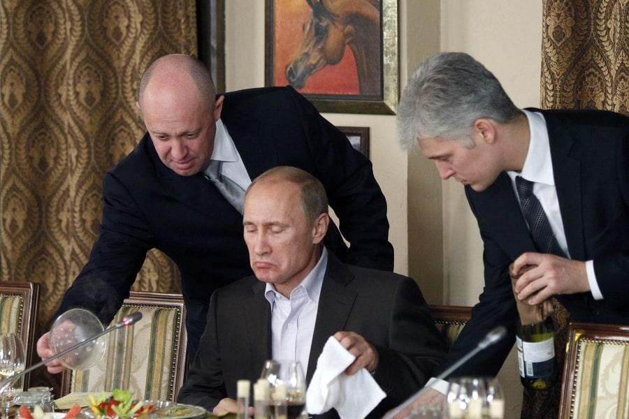 Zdjęcie z Kremla. Władimir Putin siedzi przy stole. Nad nim Jewgienij Prigożyn podaje Putinowi jedzenie. Po prawej stronie siwy mężczyzna z butelką wina.