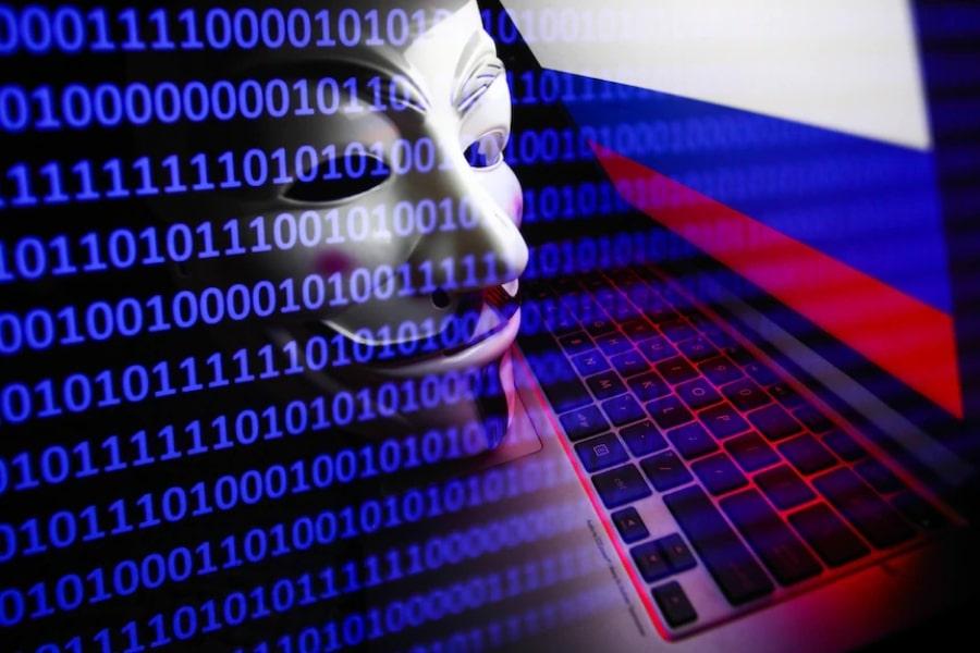 Maska Anonymous pochyla się nad laptopem. Na ekranie flaga Rosji. Na całości nałożony filtr z systemem 0 1