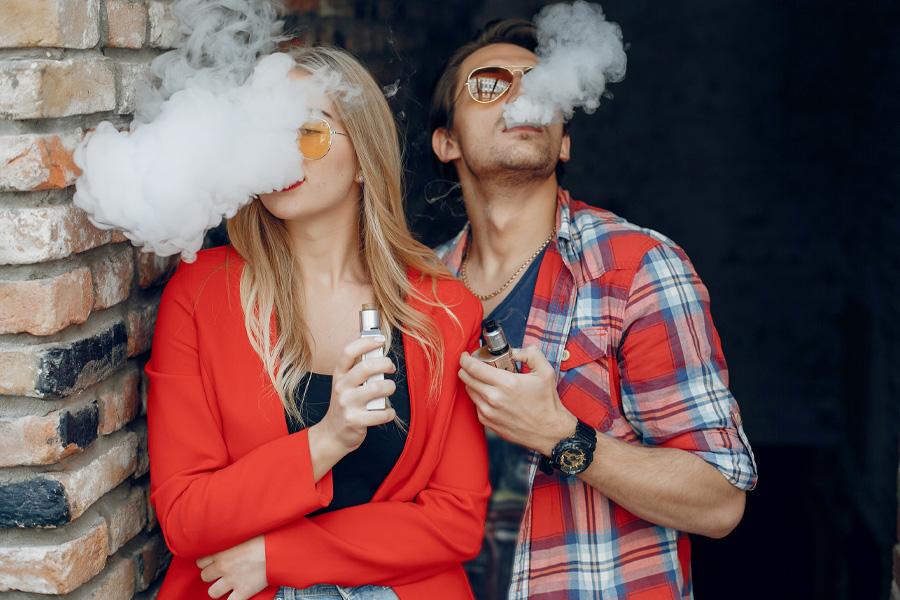 Dwójka nastolatków pali e-papierosy. Wypuszczają dym ustami.