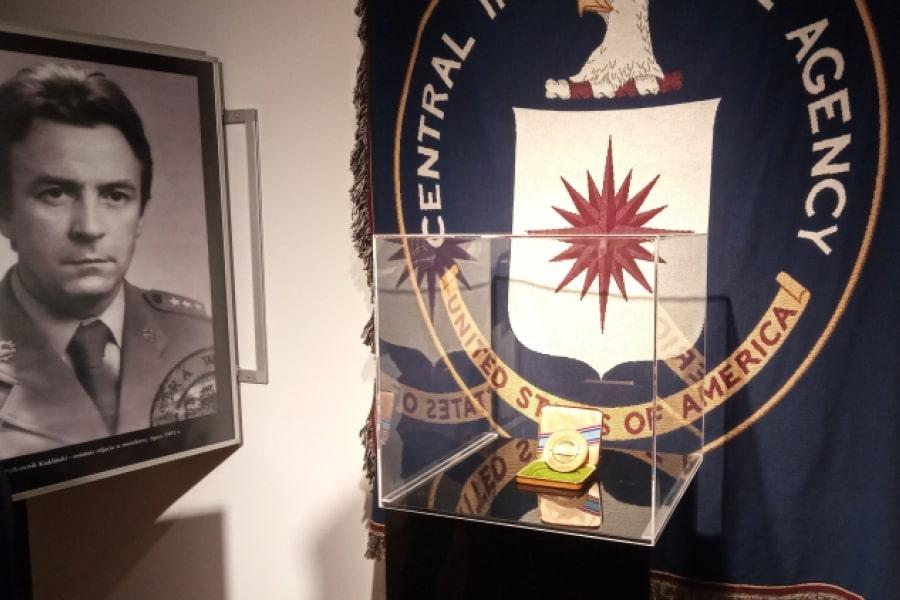 Zdjęcie pamiątkowego medalu za zasługi, który otrzymał Ryszard Kukliński. W tle sztandar CIA. Po lewej stronie czarno-biały portret Kuklińskiego oprawiony w ramie.