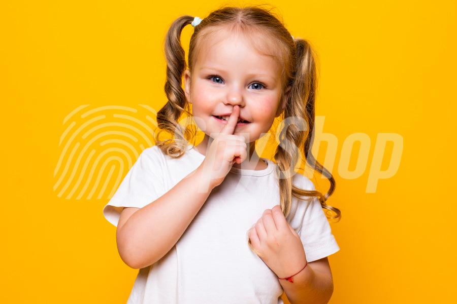 Mała dziewczynka w białej koszulce na żółtym tle. Dziecko uśmiecha się i przykłada palec do ust.
