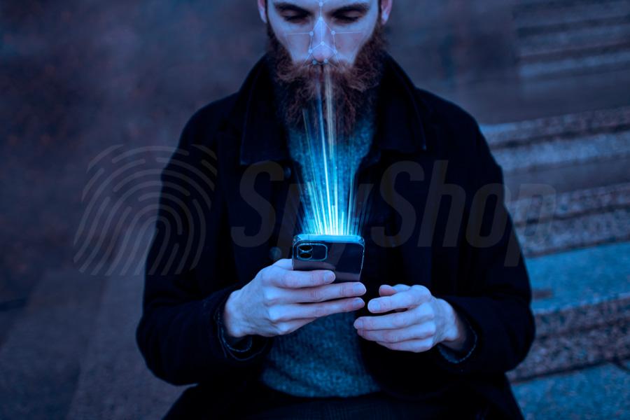 Ubrany w płaszcz mężczyzna z zarostem trzyma w dłoni smartfon. Z ekranu niesie się poświata.