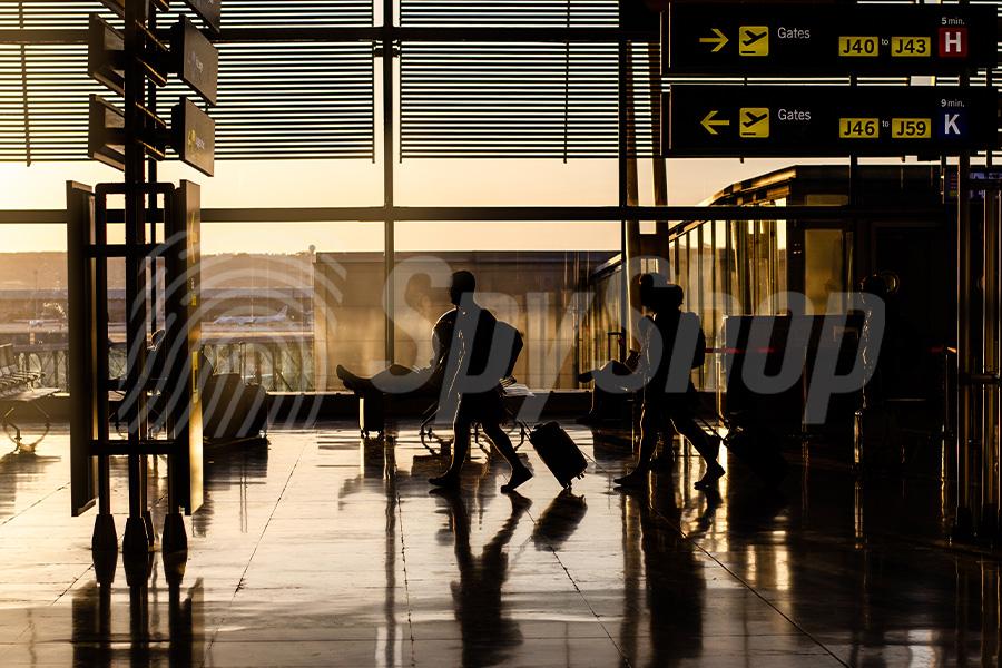 Przyciemniony przez zachodzące słońce terminal lotniskowy. Widoczni pasażerowie z walizkami, fotele oraz kierunkowskazy.