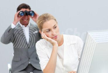 Zamyślona kobieta patrzy w ekran komputera, podpiera głowę dłonią. Za jej plecami szpieg z lornetką przyłożoną do oczu.