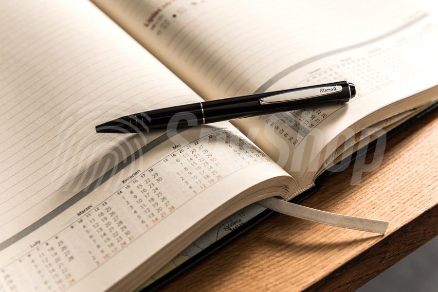 Zbliżenie na czarny długopis z dyktafonem. Przyrząd leży na otwartym kalendarzu książkowym.