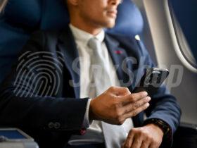 Mężczyzna w garniturze siedzi w samolocie. W prawej dłoni trzyma telefon.