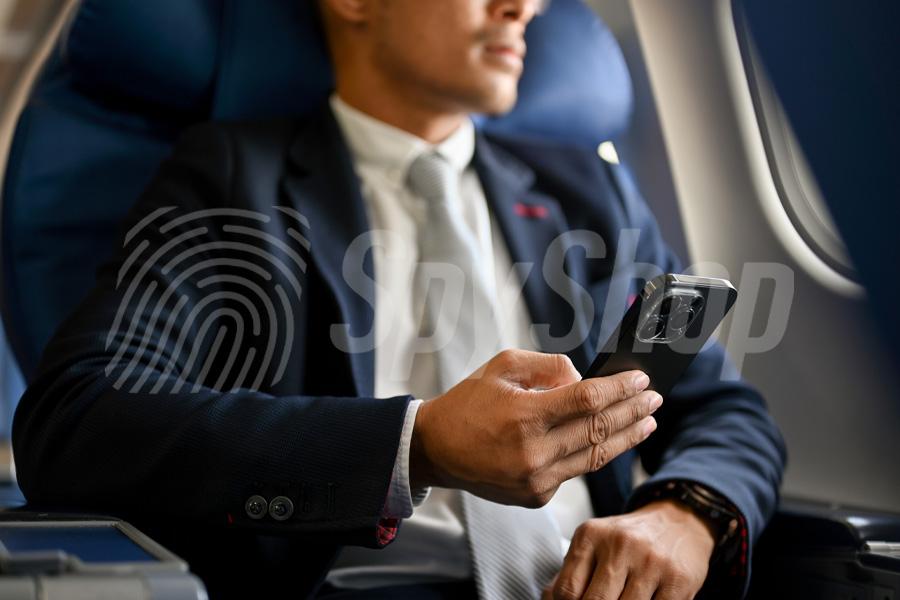 Mężczyzna w garniturze siedzi w samolocie. W prawej dłoni trzyma telefon.